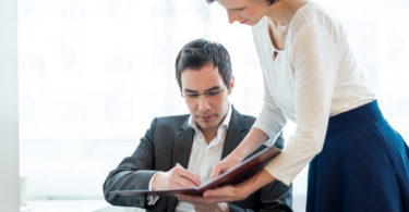 Secretary standing in the office presenting a document in a file to her boss seated at his desk for his clearance and signature.