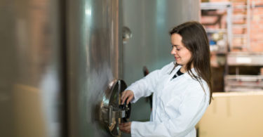 Female technologist in white overall near tub filled with olive oil in manufacturing environment