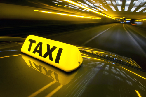 A cab at high speed on a motorway in an urban area with the lit taxi sign on top of its roof