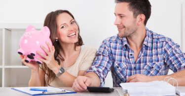 Young Man Looking At Woman Inserting Coin In Piggybank