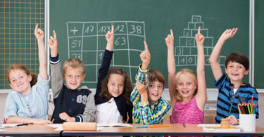 Enthusiastic group of young kids in class sitting in a row at their desk raising their hands in the air to show the know the answer to a question