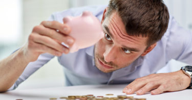 business, people, finances, crisis and money saving concept - businessman shaking piggy bank and getting coins at office