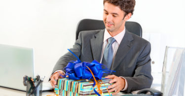 Pleased modern businessman sitting at office desk and looking on gift