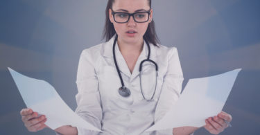 Nurse in white robe and black glasses is writing her report and thinking about it at the  table against of grey background