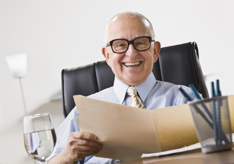 An elderly business man seated in an office and smiling.  He is holding a file folder and is wearing glasses.  Horizontally framed shot.