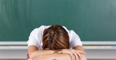 Teacher suffering from acute stress resting her head on her arms at her desk in front of the blackboard as she seeks to gather herself together