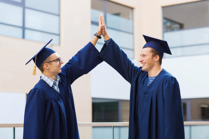 education, graduation and people concept - smiling students in mortarboards and gowns making high five gesture outdoors