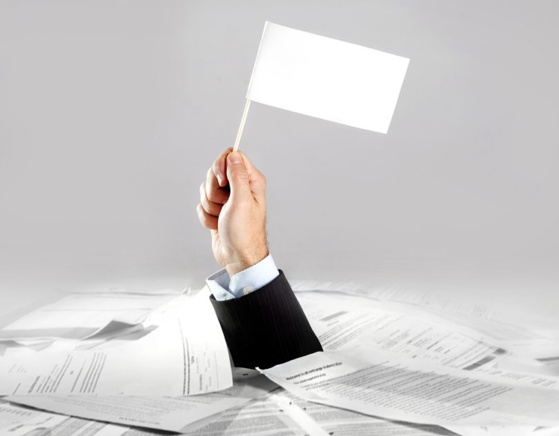 Hand of caucasian businessman emerging from office desk loaded of paperwork , invoices and a lot of papers and documents holding white flag  overworked in giving up concept