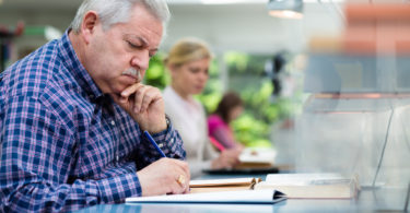 Elderly man studying with a group of young college students in library and taking notes