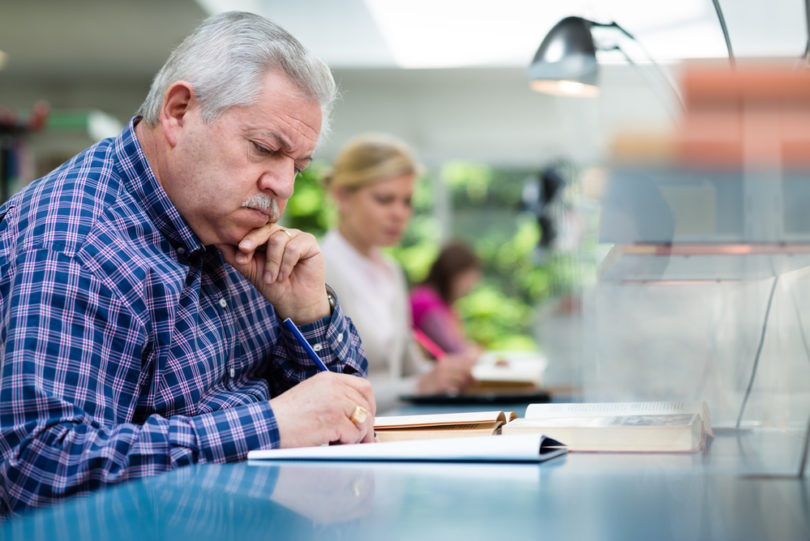 Elderly man studying with a group of young college students in library and taking notes