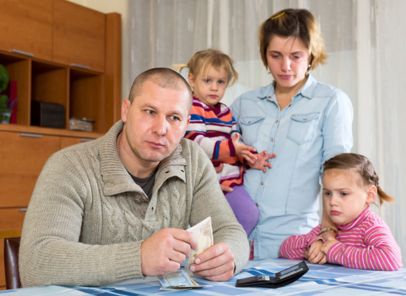 Family having problems with cash. Father is sitting at the table with money in his hands his wife and two sad daughters are sitting behind him